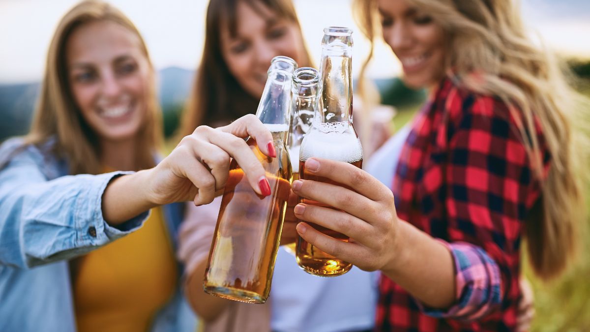 Alcool et organisme jeune : risques et faits essentiels Trois jeunes femmes trinquent avec des bouteilles de bière en plein air.