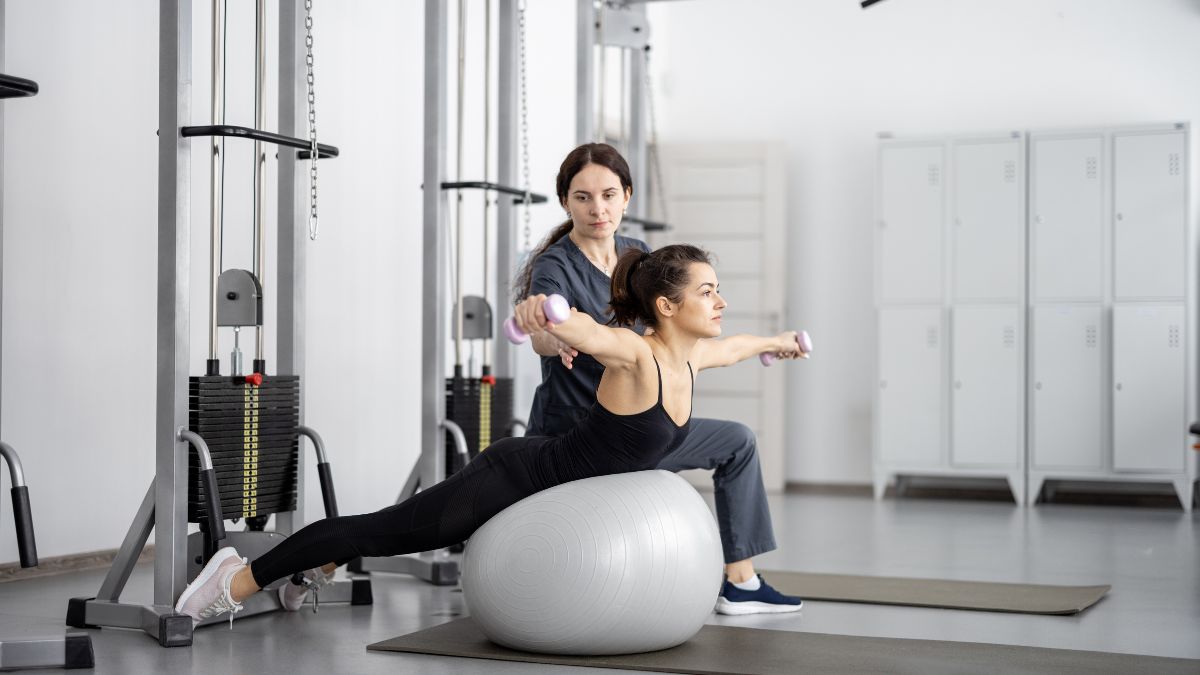 Une femme s'exerçant sur un ballon de gym avec des haltères sous la supervision d'une coach.
