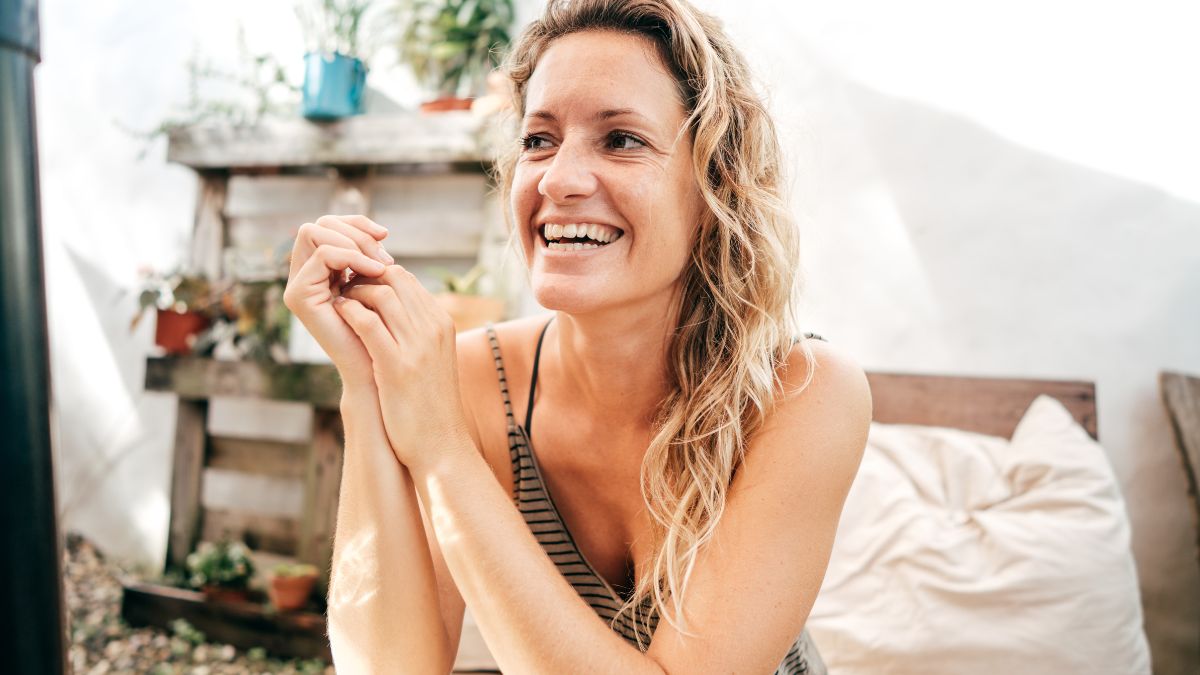 Femme souriante avec des cheveux ondulés, assise dans un environnement lumineux et naturel.